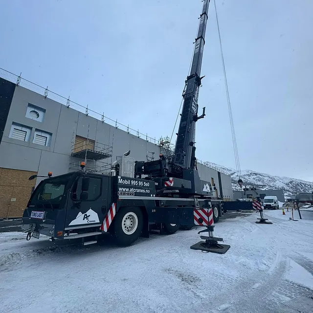 Stor mobilkran står på snødekt byggeplass foran industribygg.