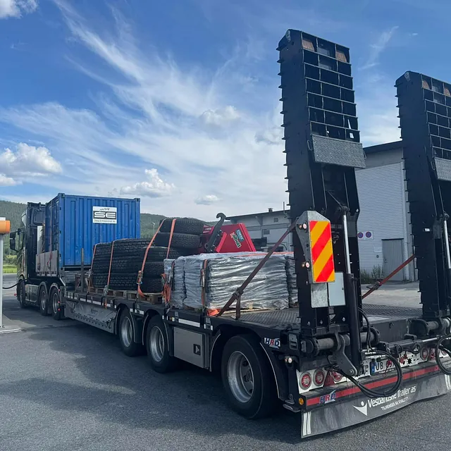 Lastebil med henger frakter dekk og blå container ved bensinstasjon.