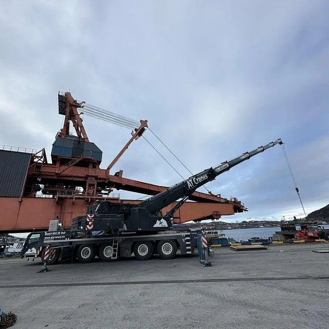 Mobilkran står ved kaiområde foran store lastekraner og sjø.
