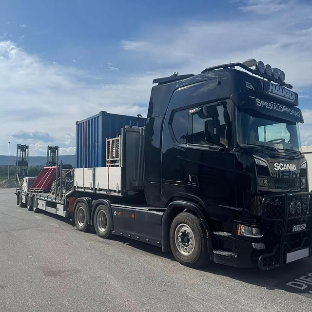 Svart lastebil med henger frakter container og utstyr på åpen vei.