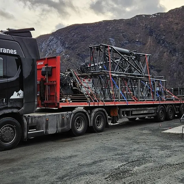 Lastebil med henger frakter store krandeler foran fjell og verkstedhall.