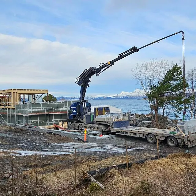 Kranbil løfter materialer på byggeplass ved sjø med fjell i bakgrunnen.