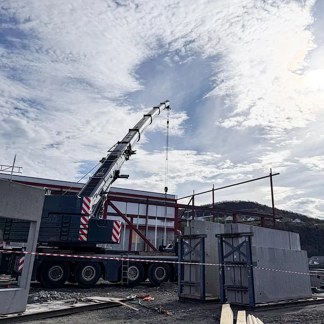Mobilkran løfter betongelementer på byggeplass under skyet himmel.